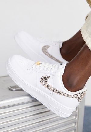 Białe sneakersy z beżowym i czarnym wzorzastym znakiem Swoosh oraz piętą, noszone przez osobę opierającą stopę na metalowej powierzchni.