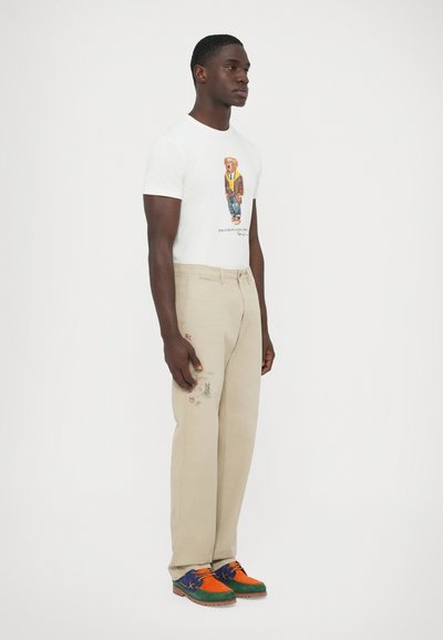 T-shirt branca com estampa de urso, calças bege com detalhes bordados, e sapatos multicoloridos com realces em azul, laranja e verde.