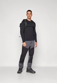 Άνδρας που στέκεται φορώντας μαύρο μακρυμάνικο πουκάμισο, γκρι και μαύρο παντελόνι, παπούτσια πεζοπορίας και μαύρο σακίδιο πλάτης, σε απλό φόντο.