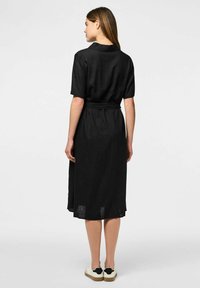 Juodas iki kelių suknelė su trumpomis rankovėmis, diržu juosmenyje ir apykaklės detale. Lygi tekstūra, krenta ties keliu, derinama su baltomis sportinėmis bateliais.