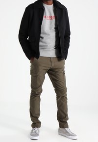 Schwarze Jacke mit Kunstpelzkragen, grauer Pullover mit rotem Logo, grüne Cargohose und graue Turnschuhe. Lässiges, geschichtetes Outfit.