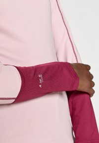 Ilgākā piedurkņu sporta tops, kuram ir rozā korpuss un kontrastējošas tumši sarkanas aproces. Materiāls šķiet gluds ar pieguļošu dizainu un logo detaļu.