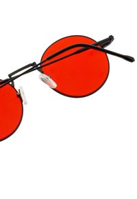 Lunettes de soleil rondes avec verres teintés en rouge, monture en métal noir et plaquettes de nez transparentes. Présente des branches noires fines et des accents matériels minimaux.
