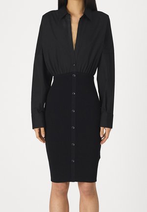 Frau trägt ein eng anliegendes schwarzes Hemdkleid mit langen Ärmeln und einem gerippten Rockteil, stehend vor einem schlichten weißen Hintergrund.