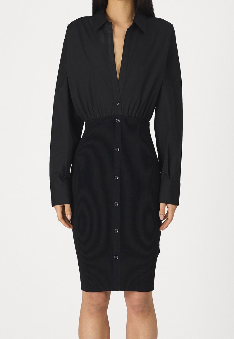 Femme portant une robe noire cintrée à boutons avec des manches longues et un panneau de jupe côtelé, debout devant un fond blanc uni.