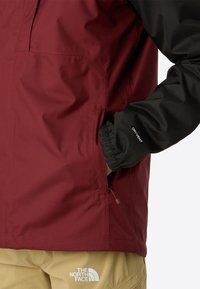 Personne portant une veste North Face rouge et noire avec une main dans une poche latérale, associée à un pantalon beige affichant le logo The North Face.