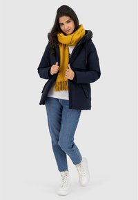 Giovane donna che indossa una giacca blu navy, una sciarpa gialla senape, jeans blu e sneakers bianche alte, in piedi con le gambe incrociate su uno sfondo chiaro.