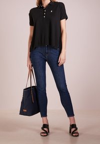 Schwarzes Poloshirt mit kurzen Ärmeln, mit Knopfleiste am Kragen, kombiniert mit dunkelblauen Jeans und einer marineblauen Tragetasche, ergänzt durch schwarze Sandalen mit Riemen.