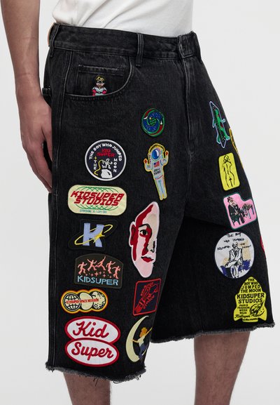 Μαύρα τζιν σορτς με πολλαπλά πολύχρωμα patches που περιλαμβάνουν θέματα διαστήματος, κείμενο "Kid Super", ένα πρόσωπο και αφηρημένα σχέδια, φορεμένα με ένα λευκό πουκάμισο.