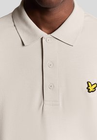 Licht beige polo shirt gemaakt van gestructureerde stof met een puntige kraag, drie knopen en een klein geel vogellogo op de borst.