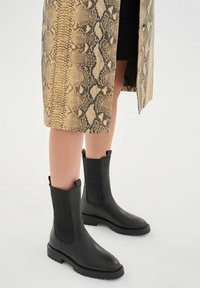 Piernas con falda de cuero de serpiente de longitud media llevando botas de cuero negras hasta la mitad de la pantorrilla con paneles laterales elásticos y tiradores sobre fondo blanco.