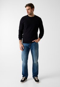 Zwarte gebreide trui met textuurpatroon, blauwe spijkerbroek met verweerd detail, en zwarte sneakers met lichte zolen. Niet-casual outfit weergegeven.