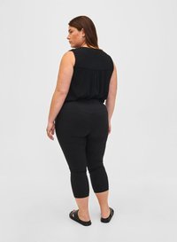 Frau mit braunen Haaren, die ein schwarzes ärmelloses Oberteil, schwarze cropped Hose und schwarze Rutschsandalen trägt, von hinten auf weißem Hintergrund gesehen.
