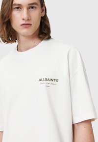 Bijela pamučna majica s kratkim rukavima i okruglim izrezom. Izvezeni tekst tamnih tonova na prsima, s natpisima "ALLSAINTS" i "LONDON."
