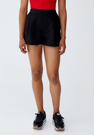 Persoon die zwarte shorts met hoge taille draagt, een rood mouwloos topje en zwarte sneakers met witte zolen, staand tegen een effen lichte achtergrond.