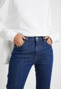 Modré džínové kalhoty s přiléhavým střihem, vybavené poutky na opasek, předními kapsami a kontrastním prošíváním. Spárováno s bílou mikinou.