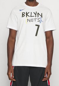 Camiseta de algodón blanca con "BKLYN" y "NETS" en negro y amarillo, número "7" en negro punteado, y logo de Nike en azul en el pecho.