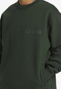 Mörkgrön sweatshirt i bomullsblandning, med rundad halsringning, långa ärmar och en framficka. Logo "Dc94" i tonalt tryck på bröstet.