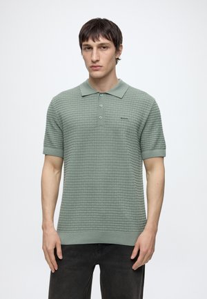 Jeune homme portant un polo à manches courtes texturé vert, debout devant un fond clair uni.