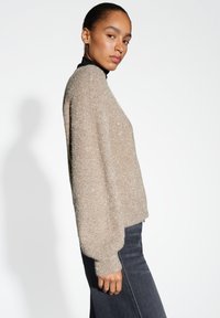 Vrouw in een sprankelende beige trui over een zwarte coltrui en donkere spijkerbroek, die zijwaarts poseert tegen een effen witte achtergrond.