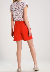 Mujer vestida con una blusa blanca de manga corta con estampado de flamencos, pantalones cortos rojos brillantes y zapatos de lona beige, de pie sobre un fondo claro.