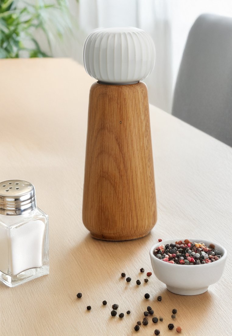Drewny młynek do pieprzu z białą górą, szklany pojemnik na sól, biała miska z mieszanką pieprzów i rozsypane kulki pieprzu na jasnym drewnianym stole.