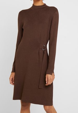 Frau trägt ein braunes, langärmliges gestricktes Kleid mit Stehkragen und seitlicher Schnallendetail, gezeigt von den Schultern bis zu den Knien.