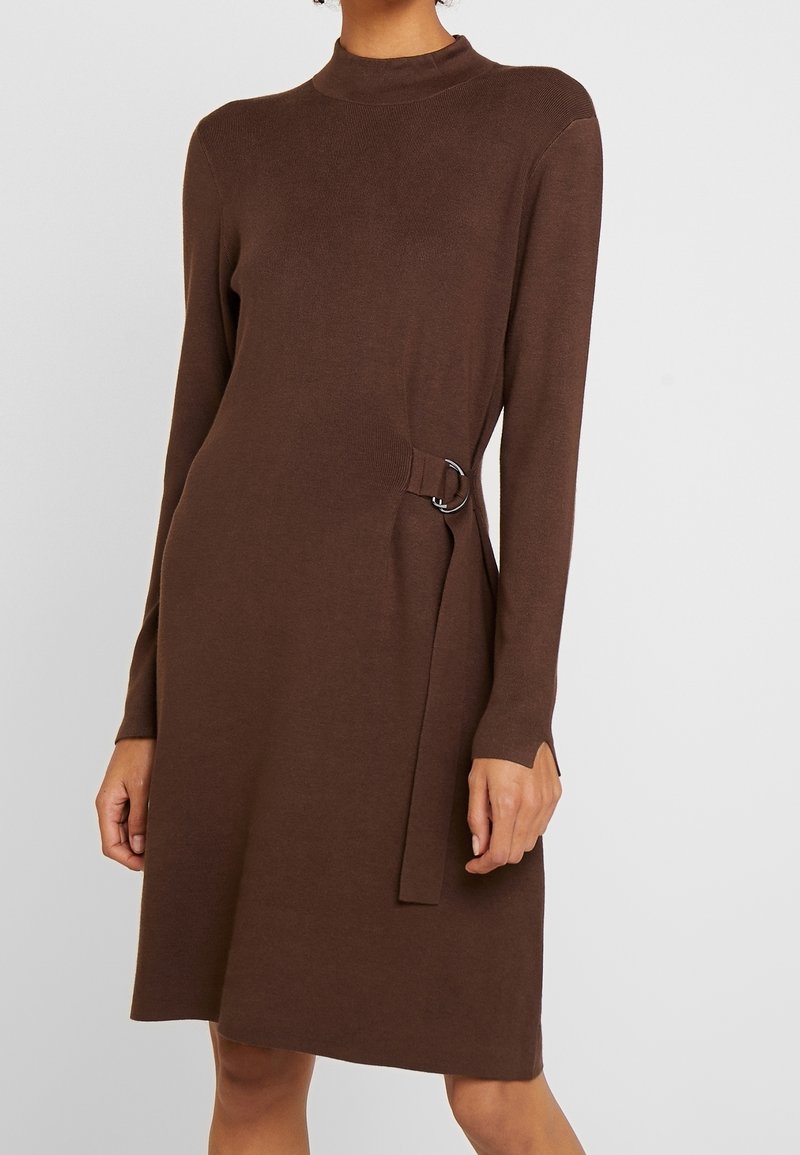 Femme portant une robe marron en maille à manches longues avec un col montant et un détail de boucle sur le côté, montrée des épaules aux genoux.