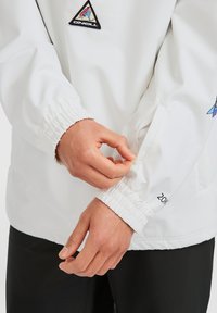 Witte jacket met elastische manchetten, een geborduurd logo-patch en verstelbare details op de mouwen. Gepersonaliseerd, lichtgewicht materiaal.