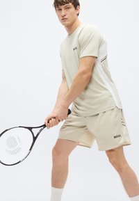 Bež športna majica in kratke hlače s črnim logotipom "BOSS". Majica ima bel panel z nežnim črtastim vzorcem. Drži teniški lopar.