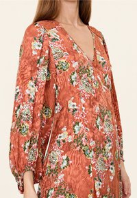 Rostfärgad klänning med blommönster, med V-ringning och puffärmar. Tyget ser ut att vara lätt och har en strukturerad yta.