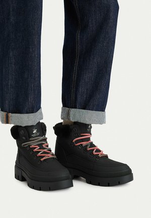 Schwarze hohe Winterstiefel mit roten und schwarzen Schnürsenkeln, Fellbesatz, getragen mit hochgekrempelten dunkelblauen Jeans auf hellem Hintergrund.