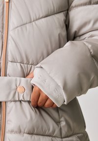 Šviesiai pilkas pūkinis striukė su užtrauktuku priekyje, pasižyminti siuvinėtomis detalėmis, diržu ir įspaustu "BAKER" ant rankovės. Papuošta rožinio aukso spaudiniu.