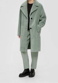 Manteau en polaire vert pâle surdimensionné avec grand col, deux boutons et poches latérales, porté sur une chemise noire et un pantalon vert clair. Bottines noires.