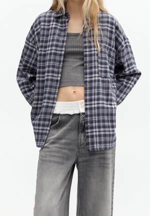 Osoba ubrana w niebiesko-białą koszulę w kratę na szarym prążkowanym krótkim topie i wyblakłych czarnych dżinsach z widocznym białym paskiem w pasie.