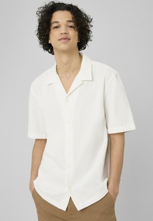 Jeune personne aux cheveux bouclés portant une chemise blanche à manches courtes boutonnée et un pantalon marron clair, mains dans les poches, devant un fond uni.