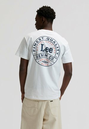 Camiseta blanca de algodón con un diseño gráfico redondo en la parte trasera que incluye "Lee Denim Co" en azul y naranja. Combinada con pantalones texturizados beige claro.