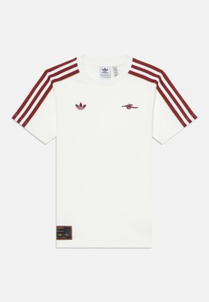 Biele tričko s krátkym rukávom, so slivkovými pruhmi na ramenách, logom Adidas, emblémom Arsenalu a čiernou etiketou na spodku. Bavlnený materiál.