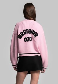 Giacca bomber lavorata a maglia rosa con texture a coste, caratterizzata da lettering nero "WRSTBHVR 030" sulla schiena e dettagli a righe nere sull'orlo.