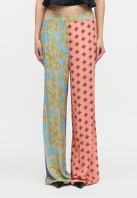 Γυναίκα που φοράει παντελόνι με φαρδιά πόδια και διαχωρισμένο σχέδιο: μπλε και χρυσό μοτίβο παϊένσια στο αριστερό πόδι, ροζ με κόκκινα διακοσμητικά διαμαντένια στο δεξί πόδι.