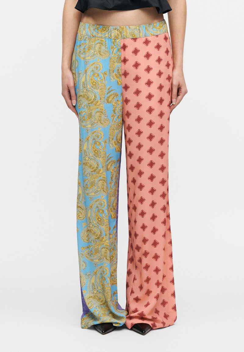 Γυναίκα που φοράει παντελόνι με φαρδιά πόδια και διαχωρισμένο σχέδιο: μπλε και χρυσό μοτίβο παϊένσια στο αριστερό πόδι, ροζ με κόκκινα διακοσμητικά διαμαντένια στο δεξί πόδι.
