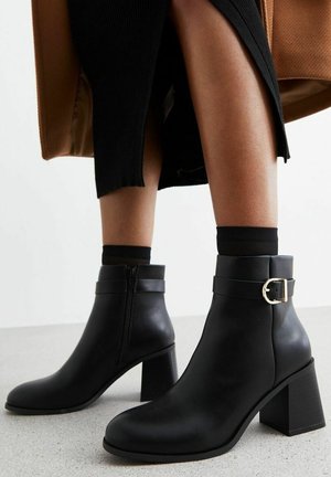 Bottes en cuir noir à la cheville avec un talon épais, dotées d'une boucle décorative et d'une fermeture éclair, au design épuré et à la texture lisse.