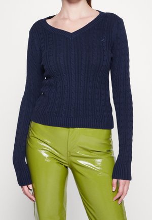 Kobieta w granatowym swetrze z dekoltem w serek o wzorze warkocza i błyszczących limonkowozielonych spodniach, widoczna od szyi do bioder na jednolitym tle.