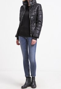 Schwarze gepolsterte Jacke mit hohem Kragen, Reißverschluss und strukturierter Oberfläche. Mit blauen Skinny Jeans und schwarzen Knöchelstiefeln kombiniert.