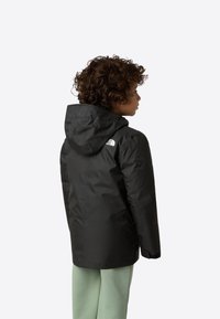 Černá voděodolná bunda s kapucí, na zadní straně s bílým logem. Hladký materiál, střední délka, v kombinaci se světle zelenými kalhotami.