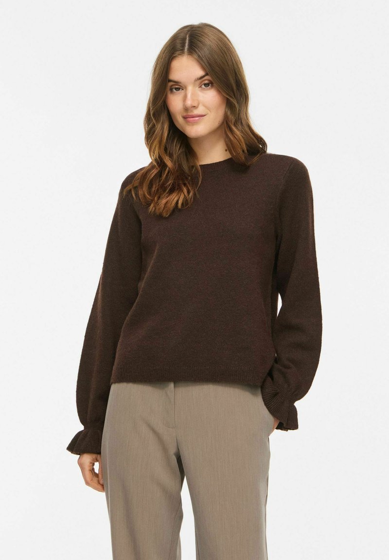 Femme aux cheveux bruns ondulés portant un pull à manches longues marron foncé et un pantalon beige, debout avec une main dans la poche sur fond blanc.
