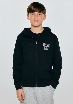 Chłopiec o ciemnobrązowych włosach, ubrany w czarną bluzę na zamek z logo oraz jasnoszare spodnie dresowe, stojący na tle jednolitego, jasnego tła.