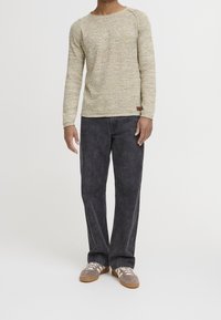 Man wearing beige long-sleeve sweater, black stonewashed jeans, and beige sneakers with white stripes, standing against a white background.