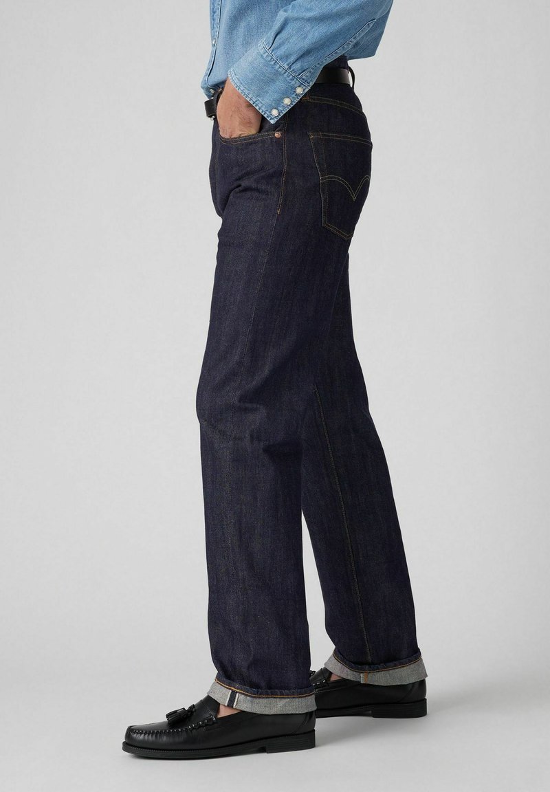 Jeans de mezclilla azul oscuro con corte recto, costuras visibles y puños enrollados. Combinados con mocasines negros y una camisa azul clara.