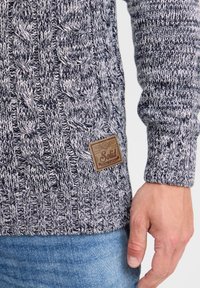 Navy and white cable-knit sweater with a textured pattern. Features a fabric label with "Solid" on the front. Worn over blue denim.